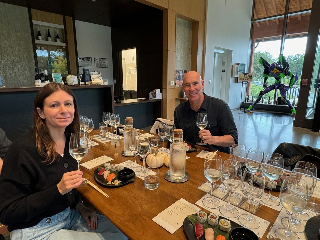 The tasting room at Dassai Blue: wine glasses of sake and plates of sushi on a wooden table, with a large anime figure standing by the windows behind