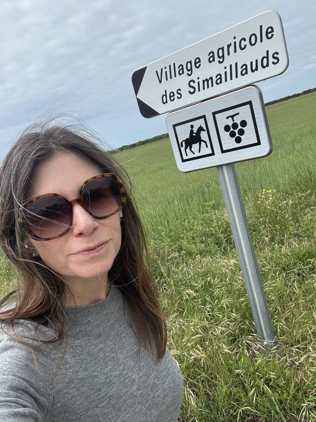 Tracey Delaney smiling beside a road sign reading Village agricole des Simaillauds, with grape and horse symbols