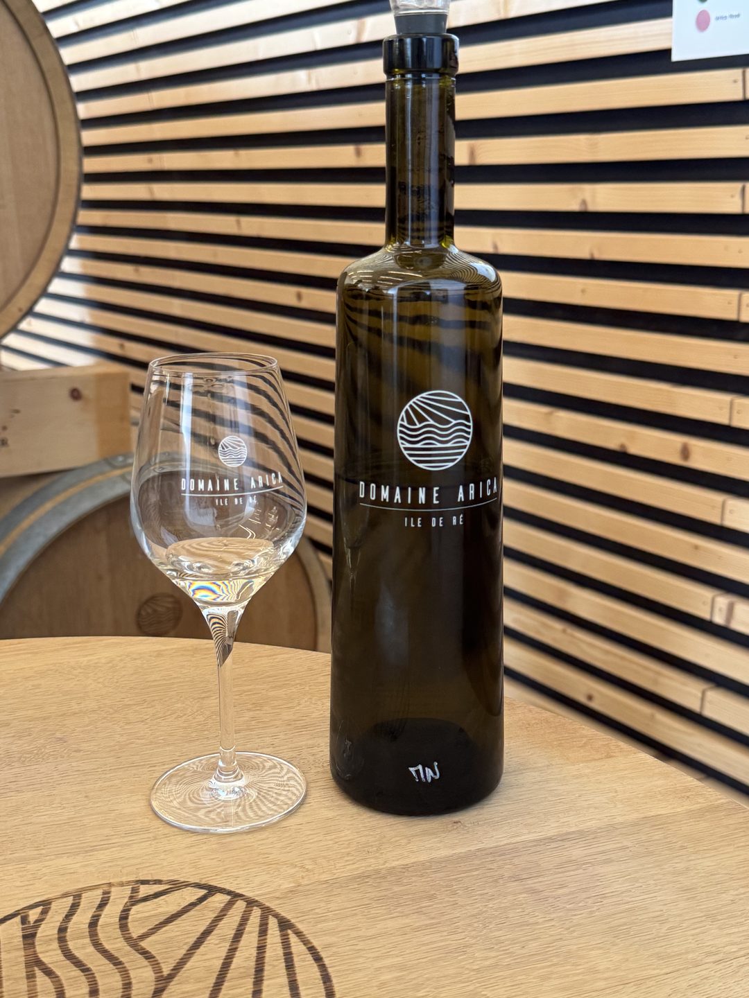 A bottle of Domaine Arica wine and a branded tasting glass on a wooden table in the tasting room, barrels behind