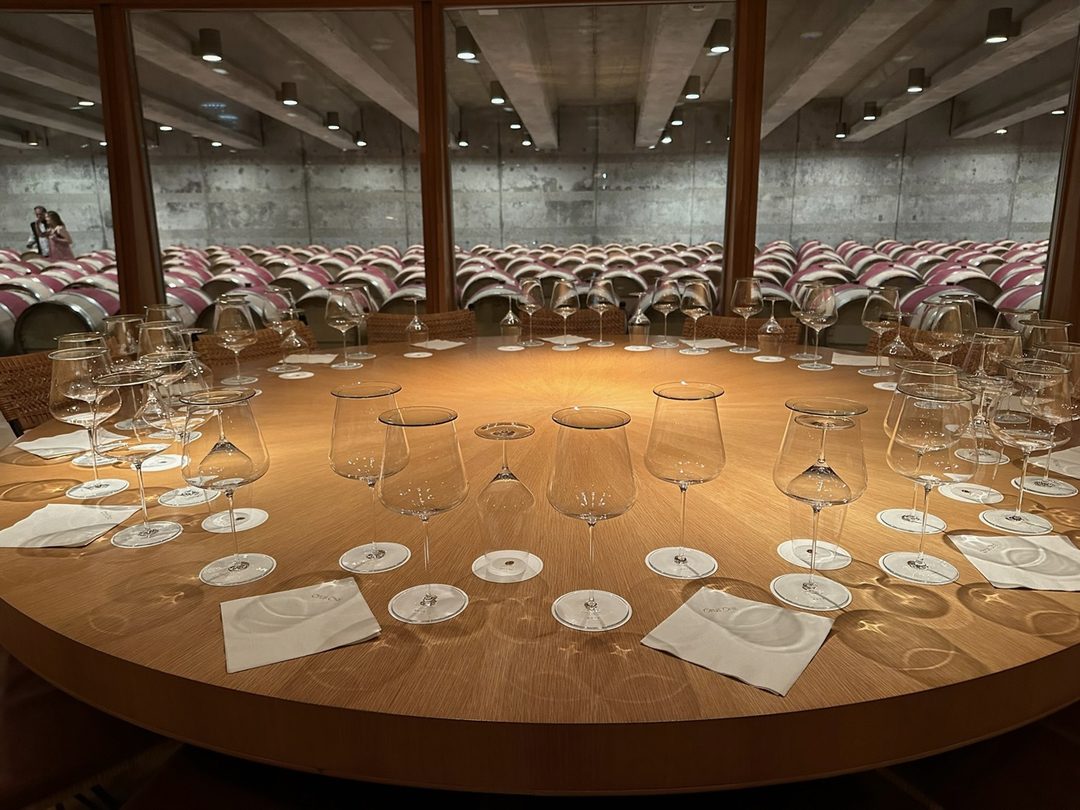 A round wooden tasting table set with wine glasses and coasters at Opus One, the barrel hall visible beyond the glass wall