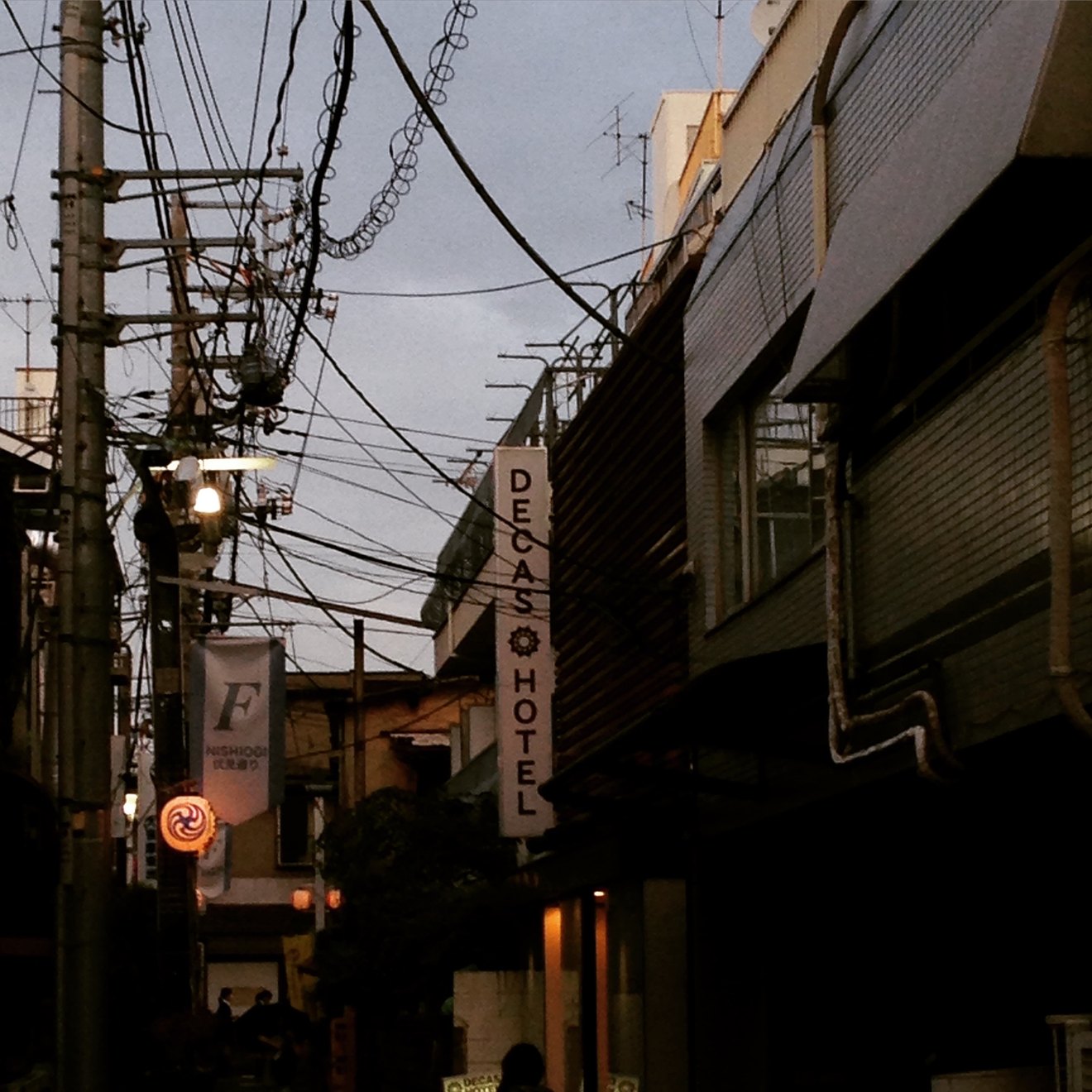 A Tokyo backstreet at dusk, shop signs lit, utility poles and wires silhouetted against the evening sky