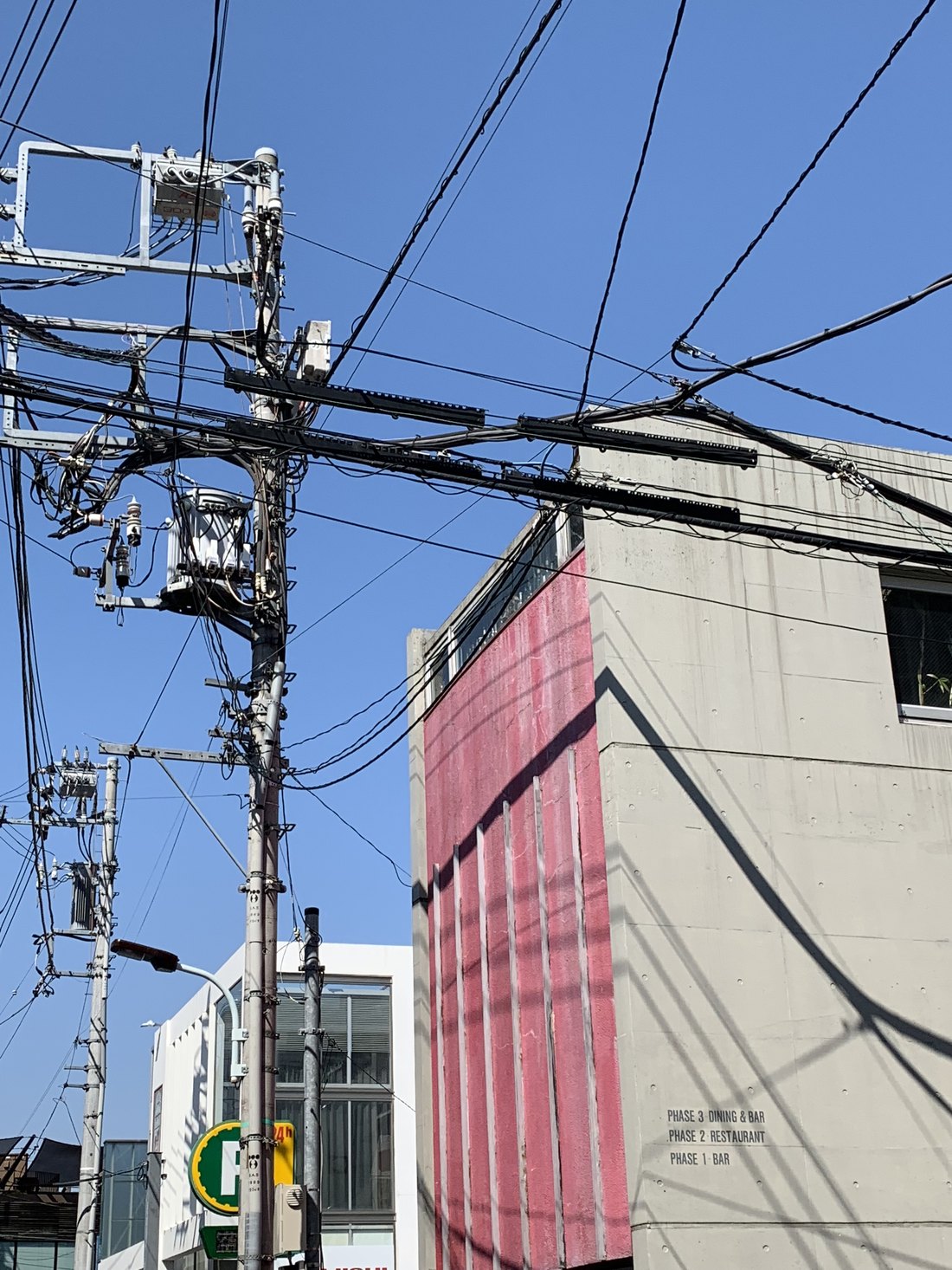 Utility poles and a dense web of wires beside a Tokyo building with a pink facade, against a clear blue sky
