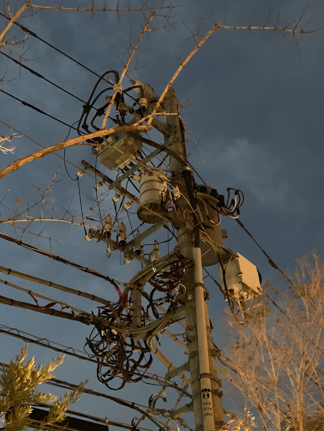 A utility pole crowded with transformers and coiled cables against a dark, golden storm sky in Tokyo