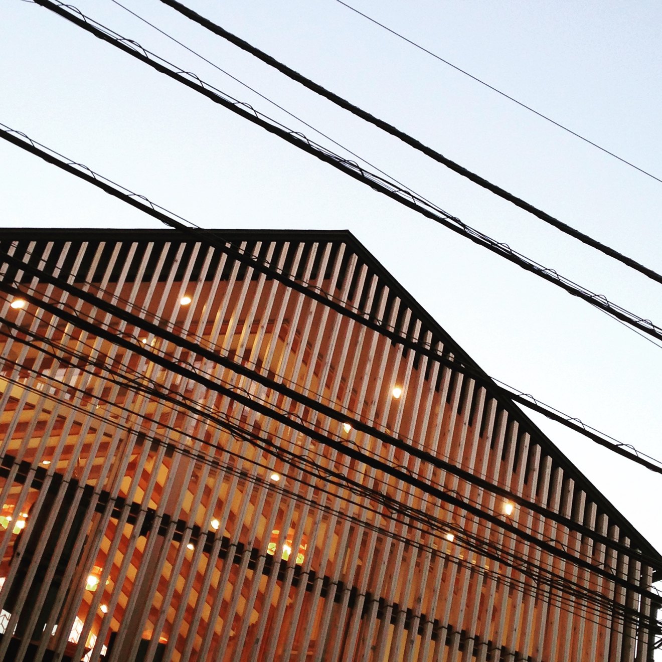 A modern wooden house in Tokyo seen through a web of overhead wires, its slatted facade lit from inside